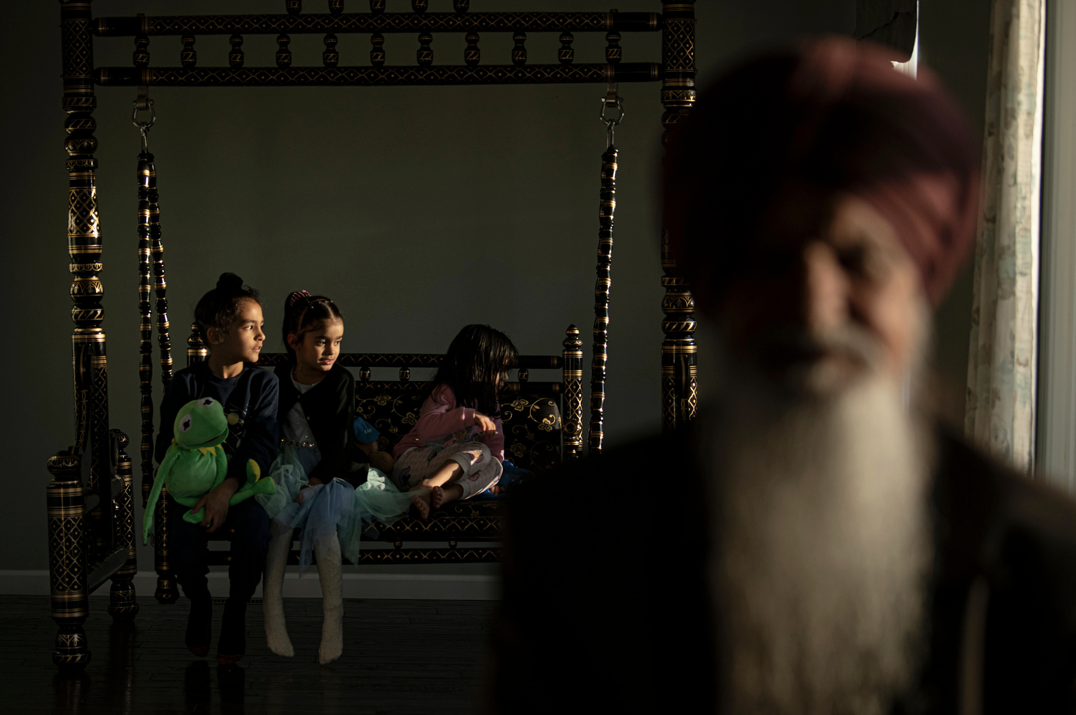 Sikh man in turban with white beard in foreground with children holding a Kermit doll behind him on an ornate bed.